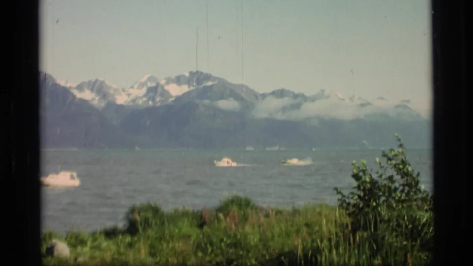 1977 Alaska 8mm frame showing boats on a fjord beyond green shoreline plants, with snow-capped mountains under hazy sky.