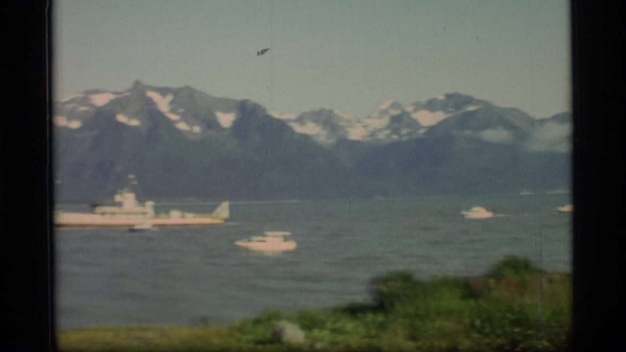 Large vessel and smaller boats on a fjord with mountains behind them.