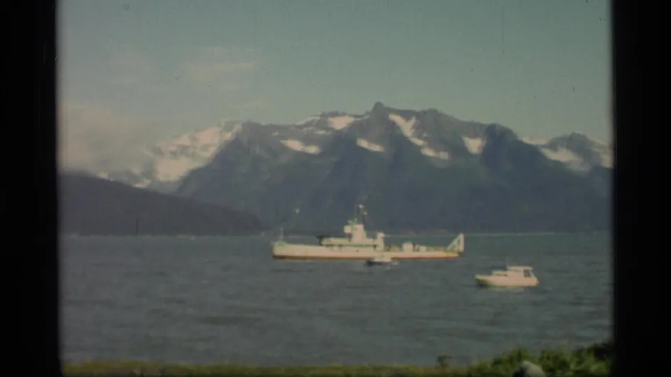 1977 Alaska 8mm frame showing a large vessel and nearby small boat on a fjord below snow-streaked mountains.