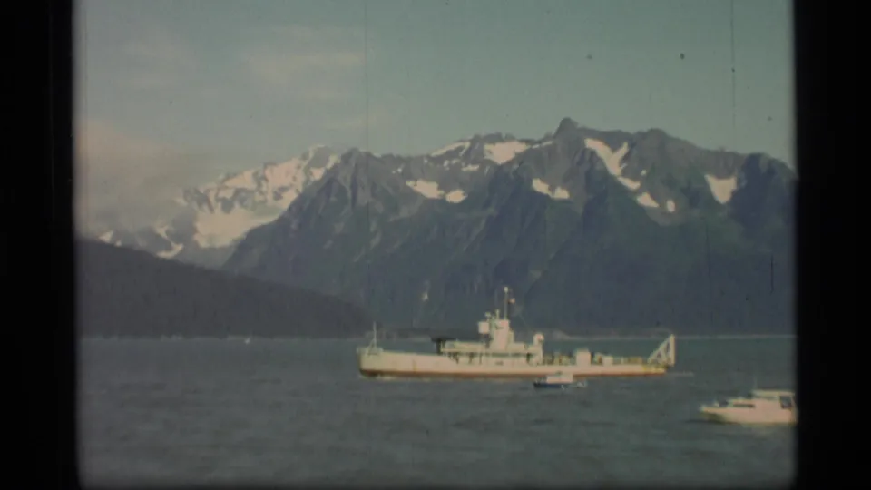 1977 Alaska 8mm frame showing a large vessel, small boats, and steep snow-capped mountains across a fjord.