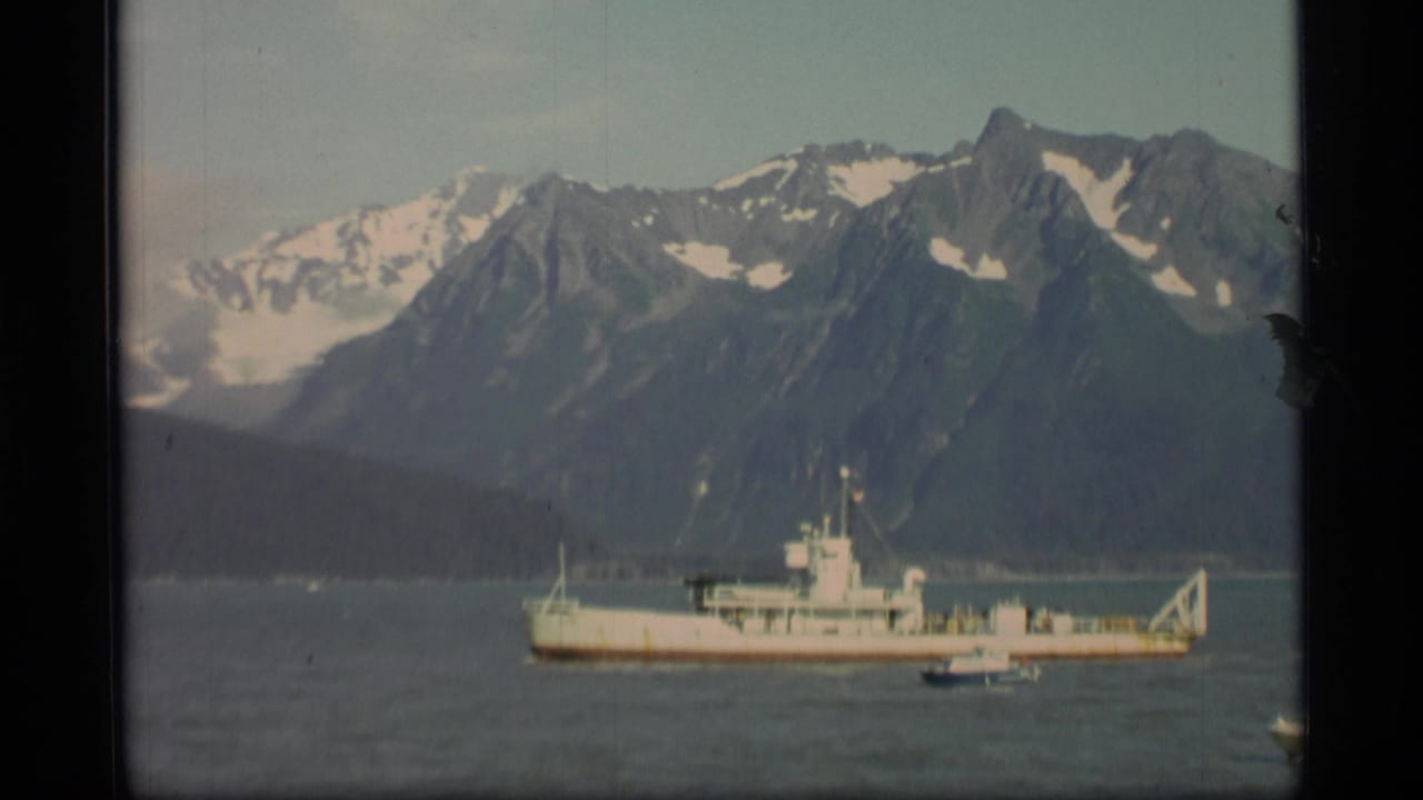 1977 Alaska 8mm frame showing a white ship crossing calm fjord water with dark shoreline and snow-capped mountains behind it.