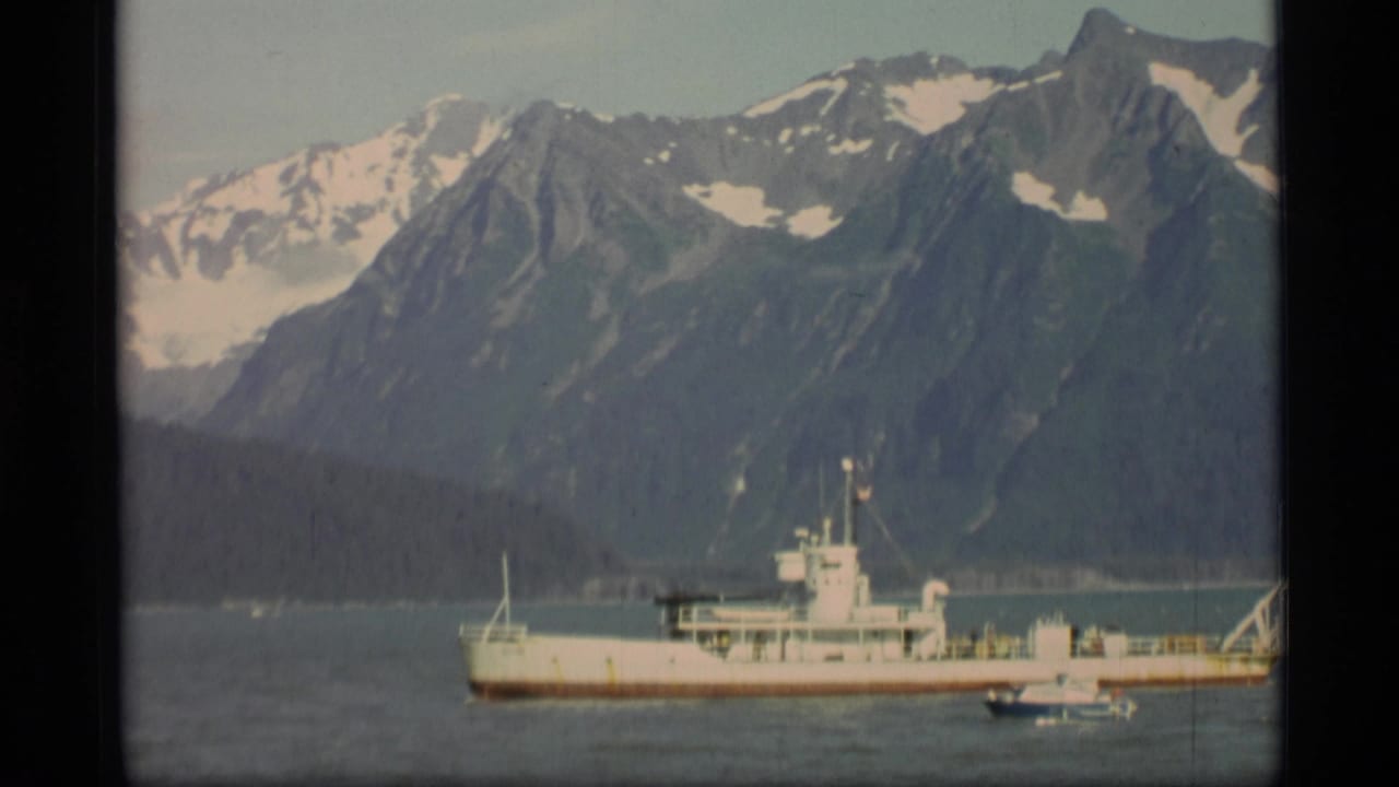 1977 Alaska home-movie frame with a white ship, nearby small boat, dark shoreline, and snow-capped mountain backdrop.