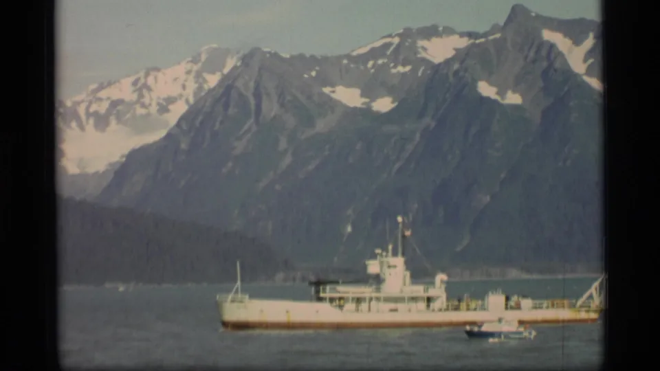 1977 Alaska home-movie frame with a white ship, nearby small boat, dark shoreline, and snow-capped mountain backdrop.