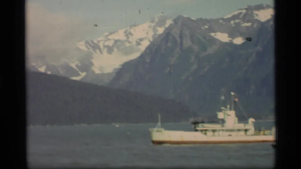 1977 Alaska 8mm frame showing the passing ship partly cropped at the right, with hazy mountains and a dark shoreline across the water.