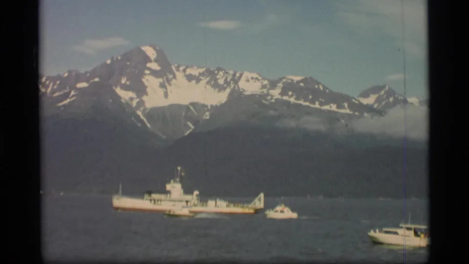 1977 Alaska 8mm frame with a distant white ship, small boats, calm water, and a broad snow-capped mountain range.
