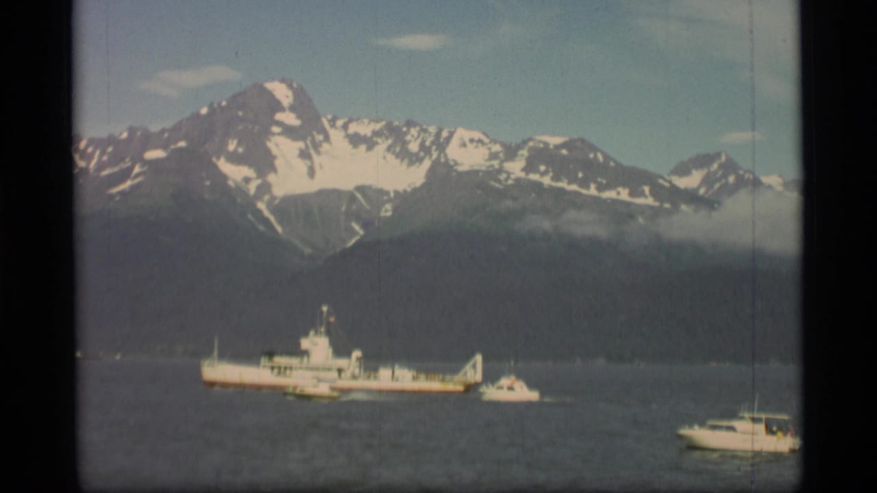 White vessel and small boats on calm water below snow-capped mountains.