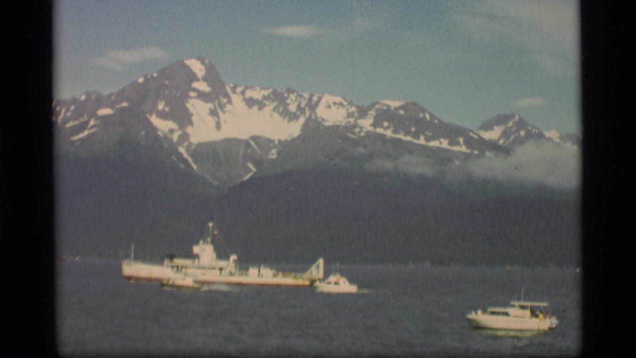 1977 Alaska home-movie frame with a white vessel offshore, small boats nearby, and a snowy mountain range in the background.