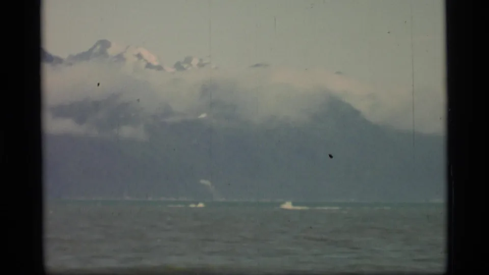 1977 Alaska 8mm frame showing low cloud over mountains and small distant boats on open coastal water.