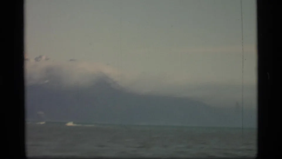 1977 Alaska home-movie frame with cloud-covered mountain slopes and calm water, with only small distant boat traces visible.