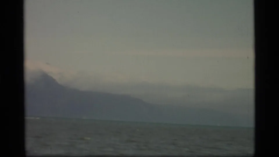 1977 Alaska 8mm frame showing gray-green ocean, hazy mountain tops, muted sky, and dark projector borders.