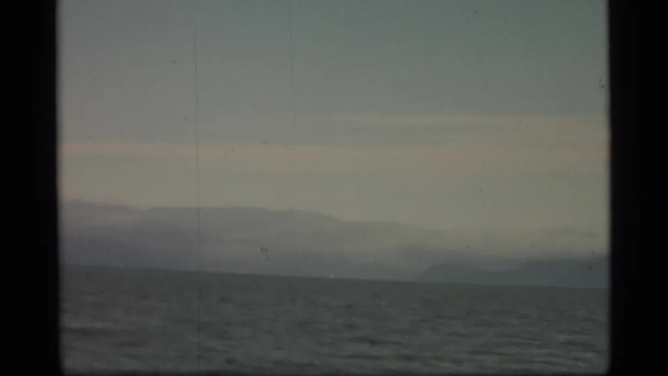 Faded 8mm Alaska coastal view with gray water, distant mountain forms, and a muted pink-gray sky band.