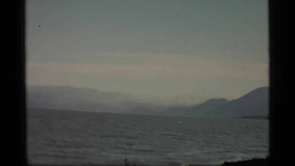 1977 Alaska 8mm frame showing choppy water, distant coastal mountains, haze, and a dark shoreline silhouette at right.