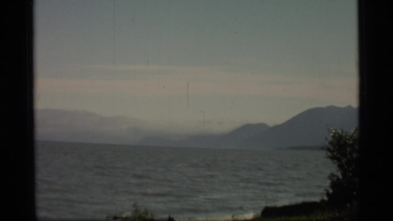 1977 Alaska 8mm frame showing calm coastal water, a hazy sky, distant mountains, and a dark foreground edge.