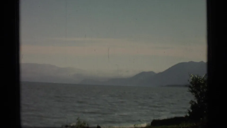 1977 Alaska 8mm frame showing calm coastal water, a hazy sky, distant mountains, and a dark foreground edge.