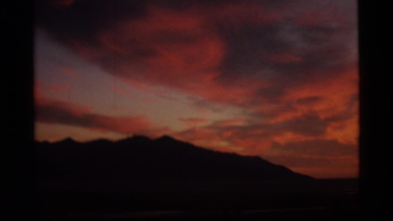 1977 Alaska 8mm frame with a dark mountain ridge against saturated red and orange sunset clouds.