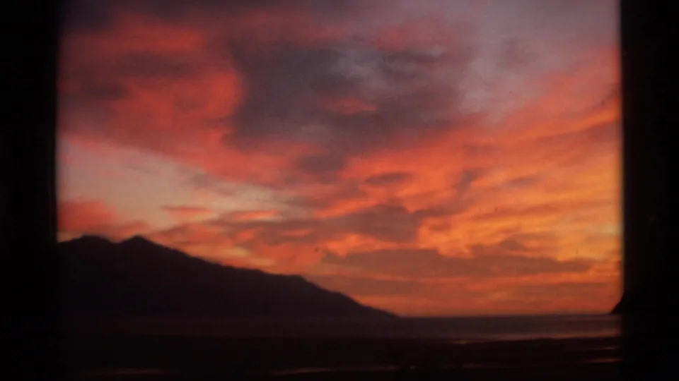 1977 Alaska 8mm frame showing a broad red-orange sunset sky above a silhouetted mountain ridge and faint waterline.