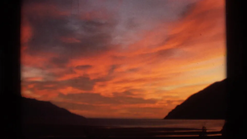 1977 Alaska 8mm frame with glowing orange clouds, dark mountain slopes, and a faint shoreline or water surface below.