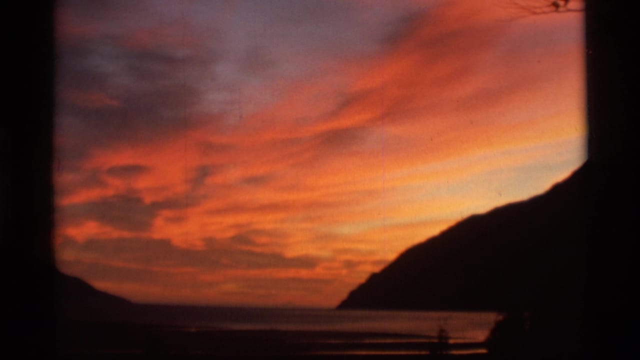 1977 8mm Alaska home-movie frame showing orange-red sunset clouds above calm sea and silhouetted coastal mountains.