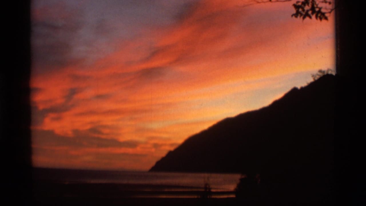 1977 Alaska 8mm frame with red-orange clouds, calm coastal water, and a dark mountain slope at the horizon.