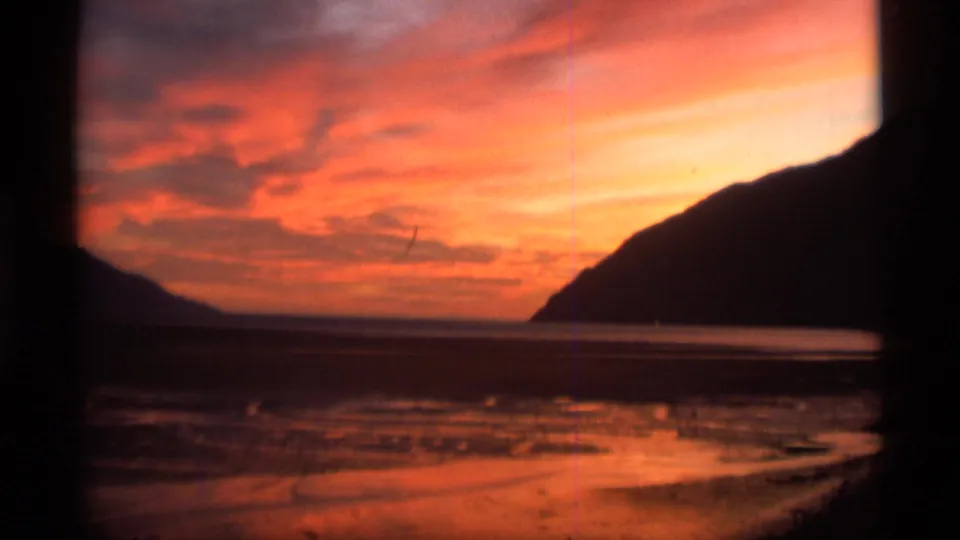 1977 8mm Alaska home-movie frame showing sunset color reflected across wet shoreline, calm water, and silhouetted coastal hills.