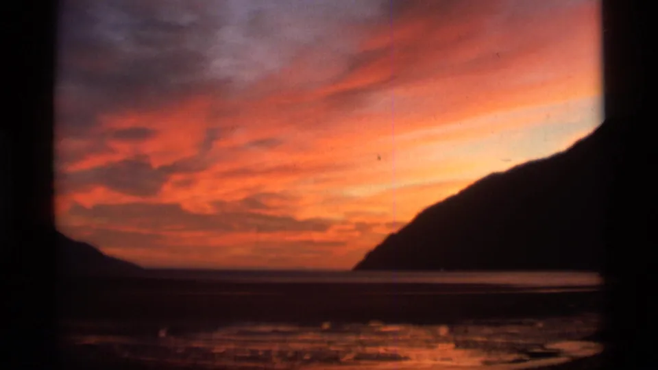 1977 Alaska 8mm frame with vivid sunset clouds, still sea, low shore, and dark mountain silhouettes at the edge of the frame.