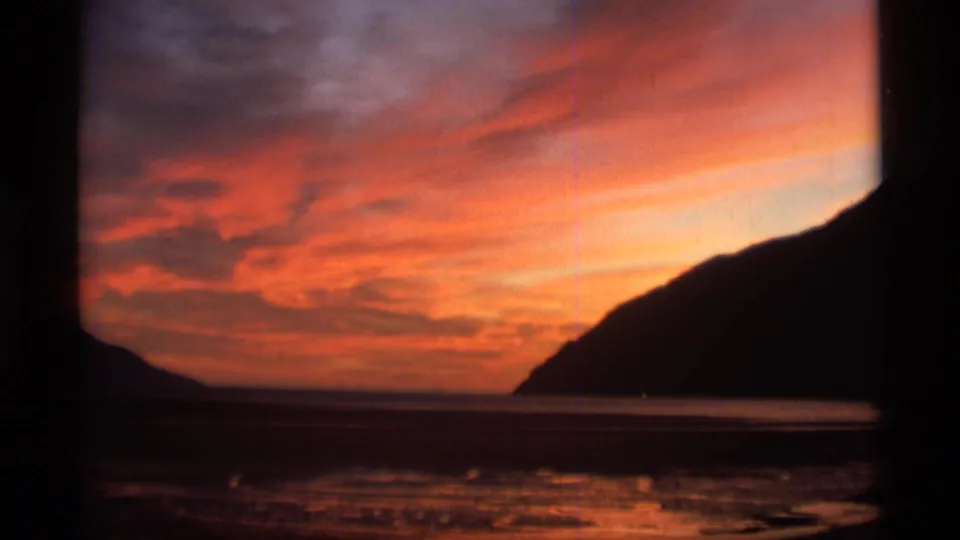 1977 Alaska home-movie frame of a red sunset over calm water beside a dark coastal mountain silhouette.