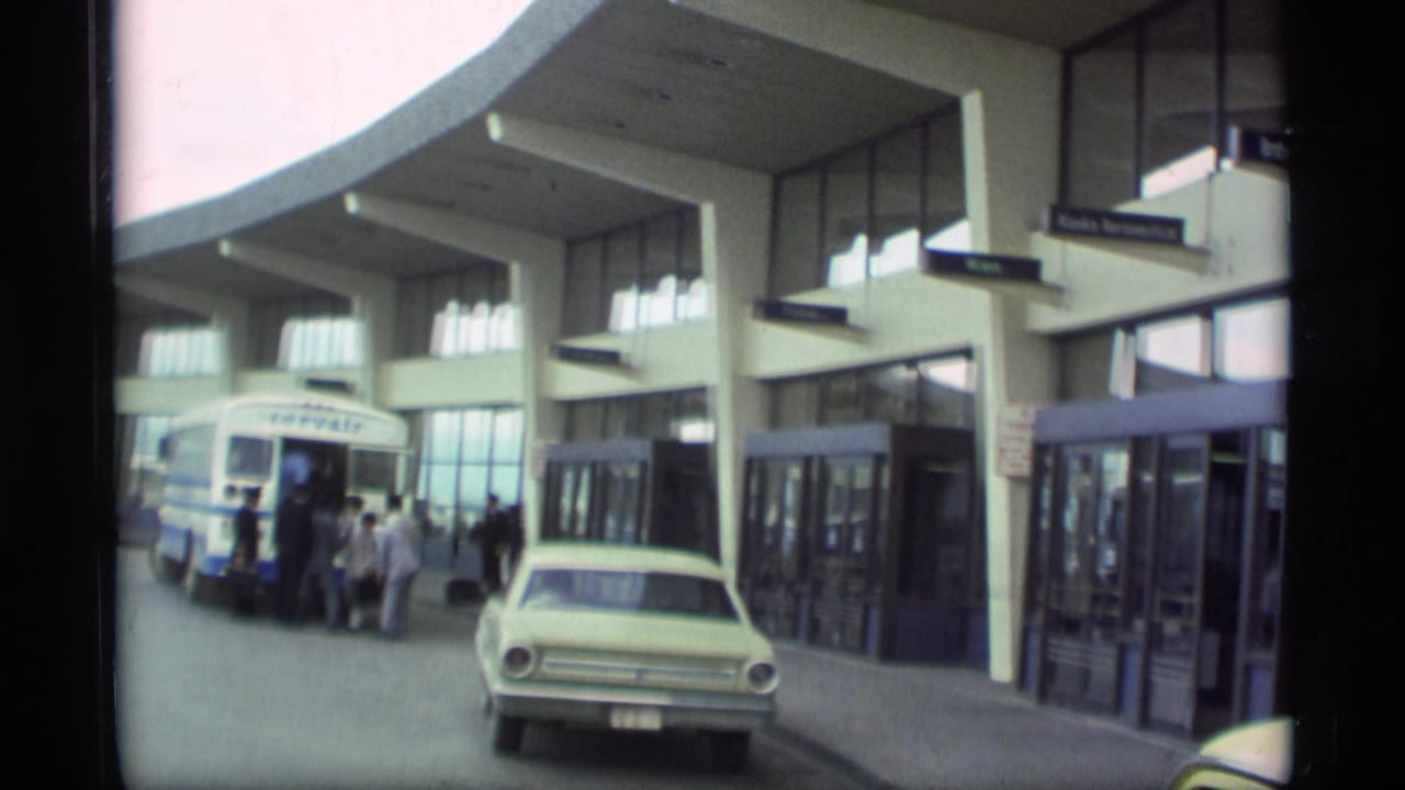 Airport terminal curbside with shuttle bus, passengers, and parked sedan.