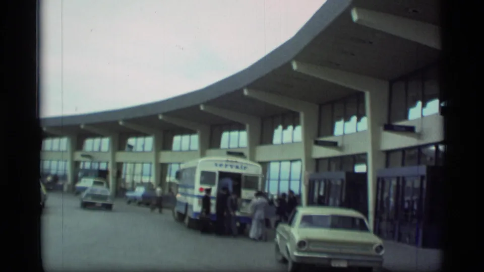 1977 Alaska airport exterior showing a curved terminal facade, shuttle bus loading area, people, and period cars.