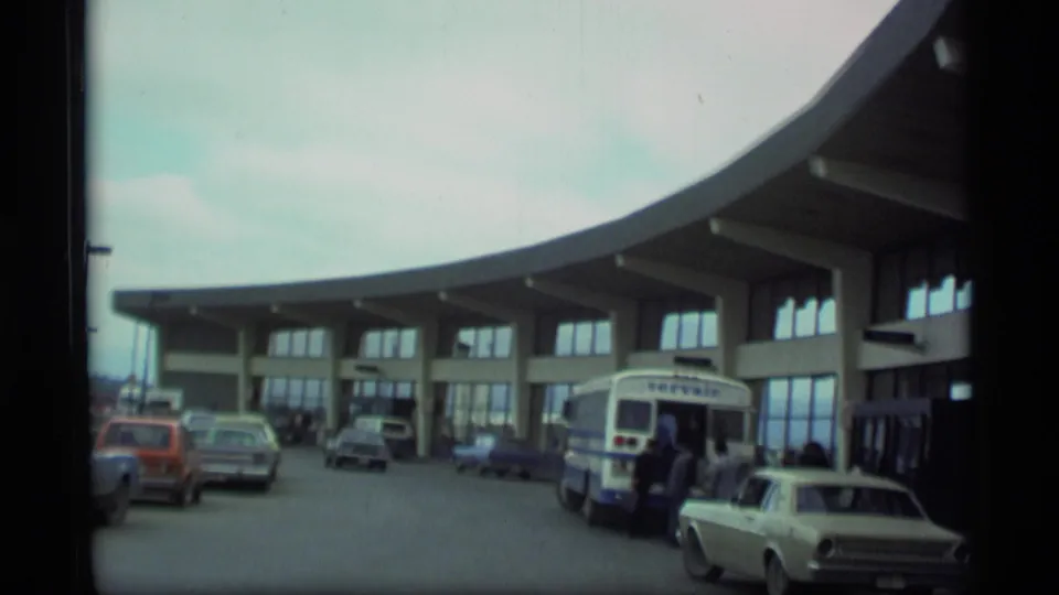 1977 Alaska airport terminal exterior with a shuttle bus at the curb, passengers nearby, and several period cars.