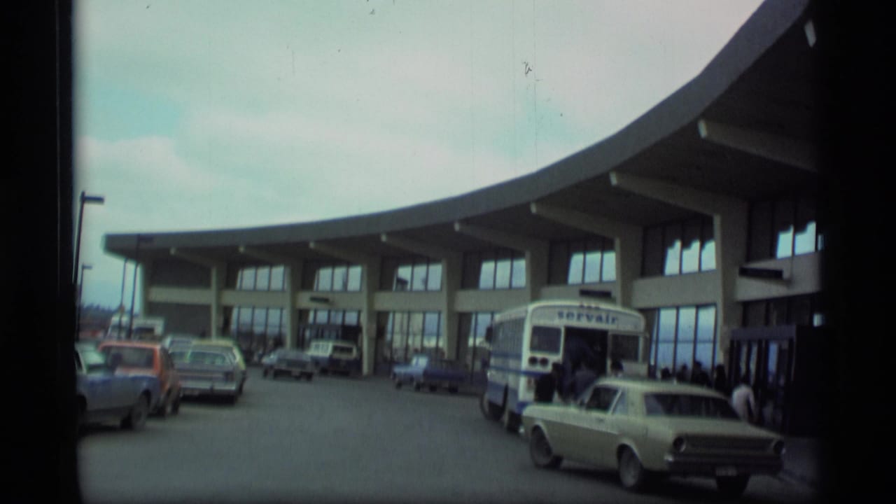 1977 Alaska airport terminal exterior with curved facade, Servair bus, parked vintage cars, and travelers near the doors.