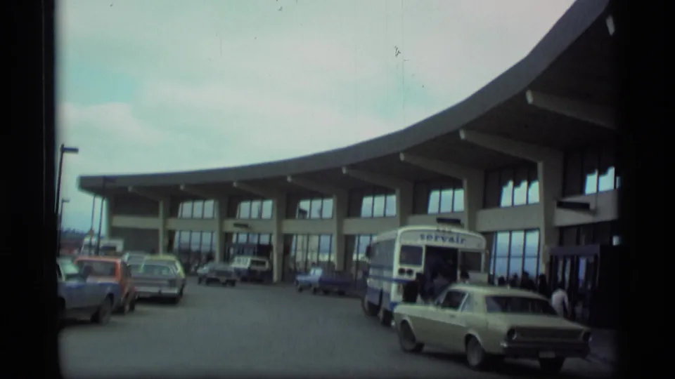 1977 Alaska airport terminal exterior with curved facade, Servair bus, parked vintage cars, and travelers near the doors.