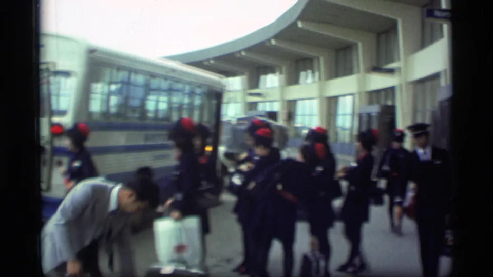 Passengers and flight attendants in dark uniforms with red hats stand beside a Servair bus outside the Alaska terminal.