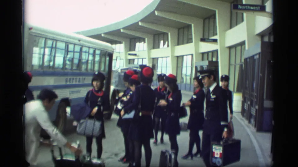 A group of flight attendants and travelers stand with bags beside the Servair bus at an Alaska airport terminal in 1977.