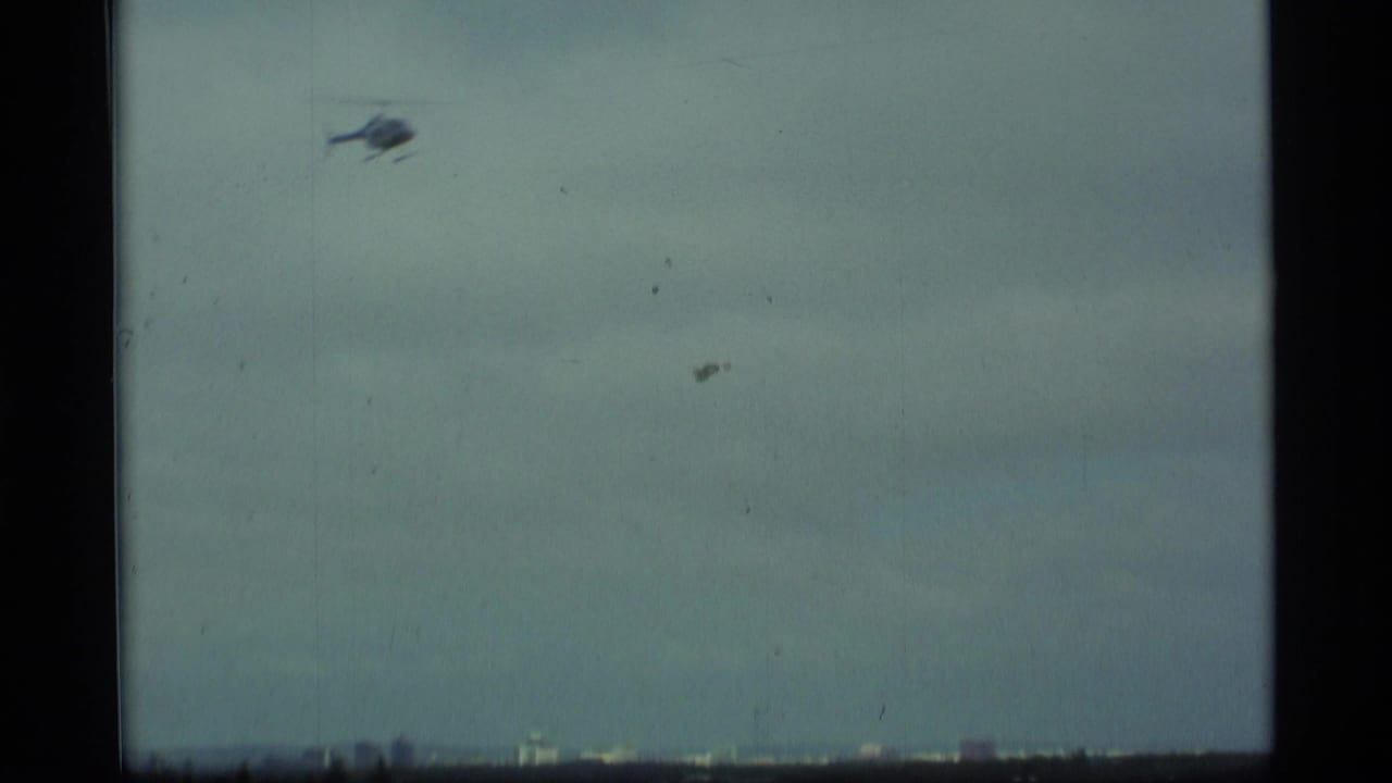 A small helicopter crosses a pale, hazy sky above a distant urban horizon, consistent with the supplied Alaska setting.