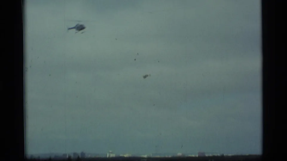 A small helicopter crosses a pale, hazy sky above a distant urban horizon, consistent with the supplied Alaska setting.