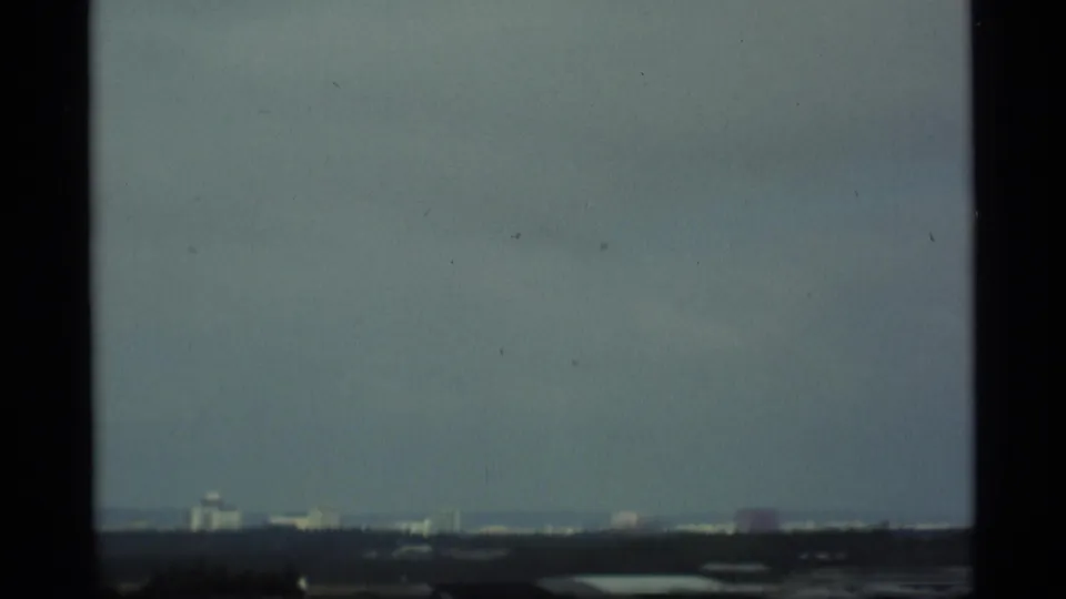 A soft 8mm view shows pale sky, a dark horizon, and small distant buildings across a hazy Alaska cityscape.