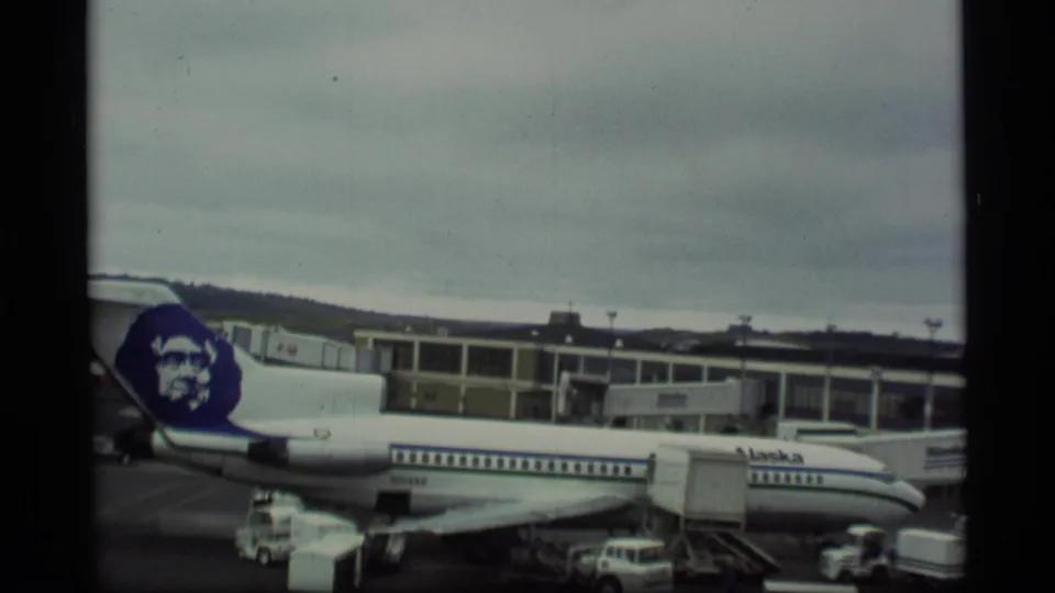 Wide view of an Alaska Airlines Boeing 727 at an airport terminal in 1977, connected to a jet bridge under gray skies.