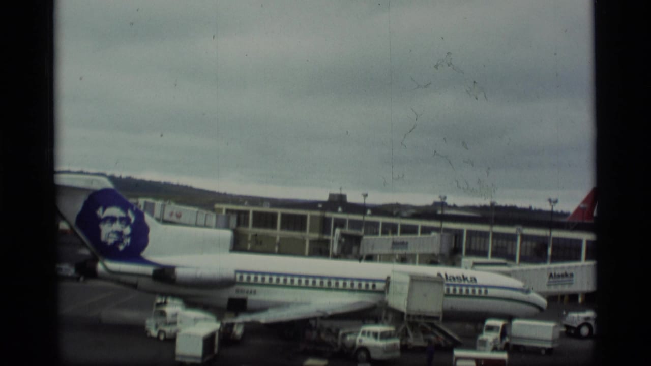 1977 Alaska travel footage showing an Alaska Airlines jet at a terminal under an overcast sky, with ground equipment and vehicles nearby.