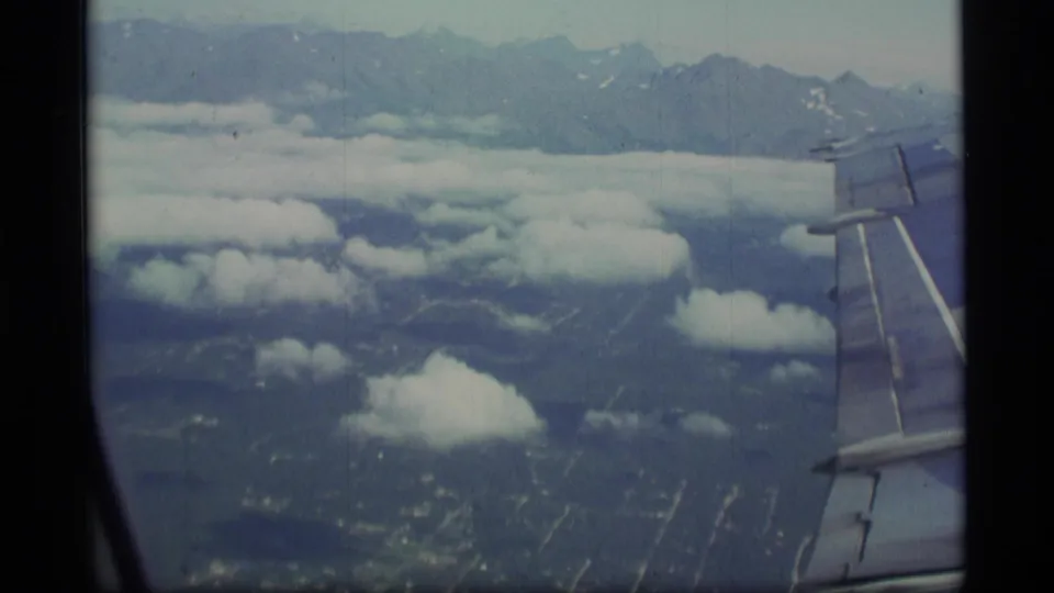 Aerial passenger-window view over clouds and distant mountains, with part of the airplane wing visible at the right edge.