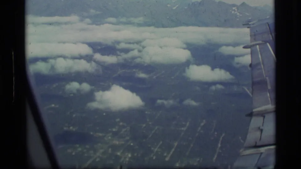 Passenger-window aerial view with the aircraft wing at right, scattered clouds, mountain slopes, and roads or city blocks below.