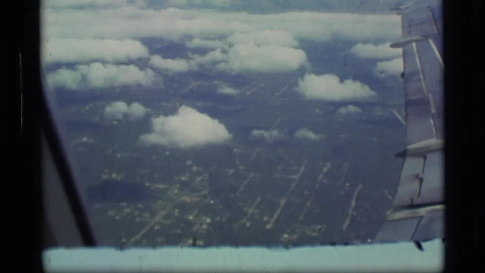 Handheld in-flight view from a passenger window, showing the wing, cloud cover, and a gridded settlement or roads below.