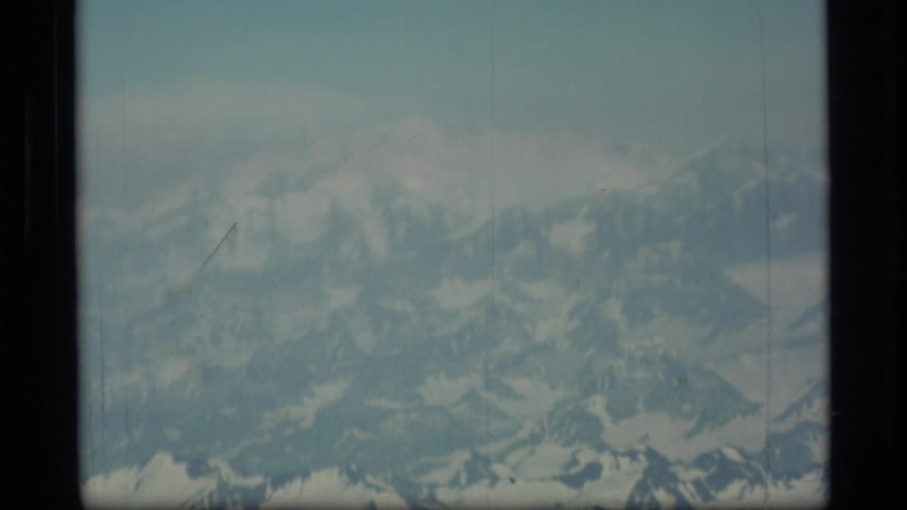 1977 8mm Alaska aerial view of distant snow-capped mountains under a pale sky, with visible film-frame borders.