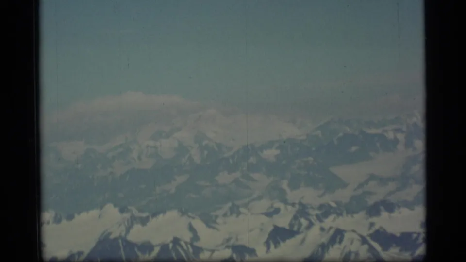 1977 Alaska home-movie frame showing an aerial view of snow-covered ridges and glaciated slopes beneath a hazy blue sky.