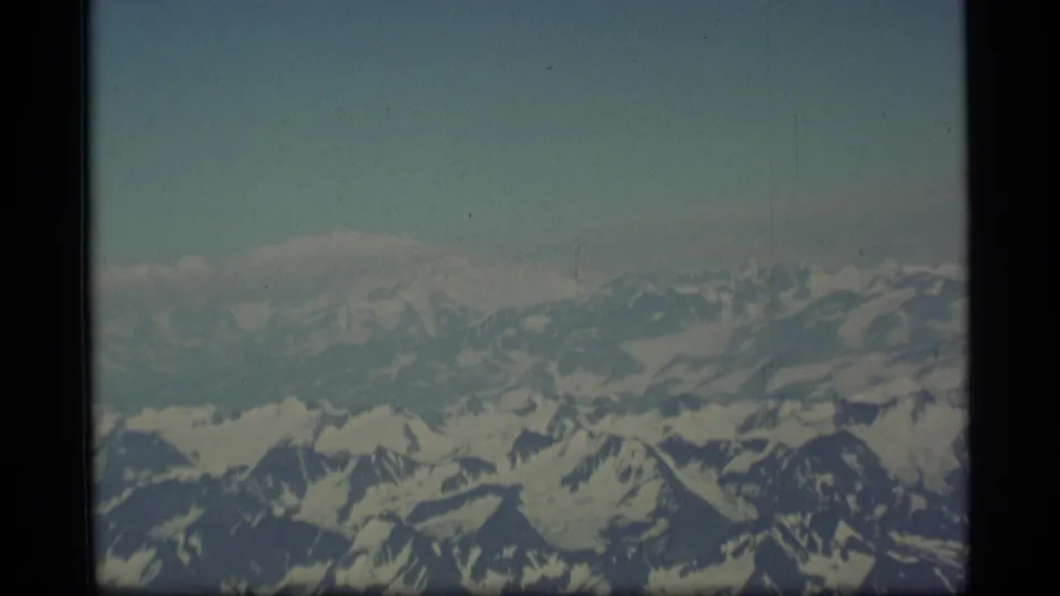 1977 8mm Alaska aerial footage frame with layered snow-covered peaks, atmospheric haze, and visible film-strip boundaries.