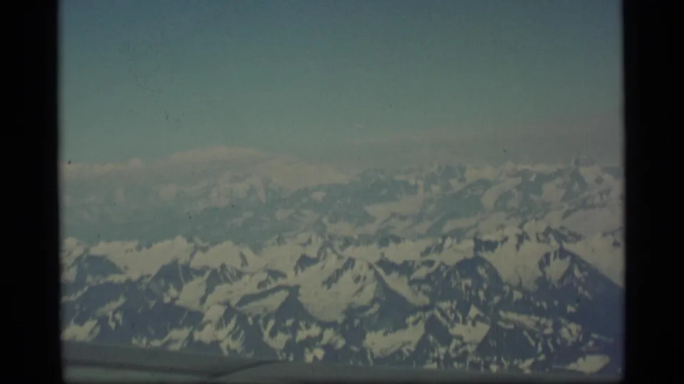 1977 Alaska aerial frame showing snow-covered mountains below a pale sky, with a dark lower edge that may be part of the aircraft window or film frame.