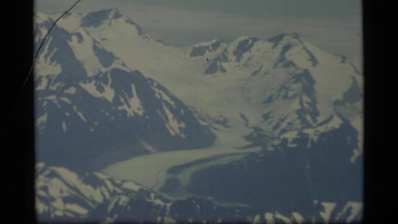 1977 Alaska 8mm aerial view of snow-covered peaks, dark ridges, and a glacier under a pale sky.