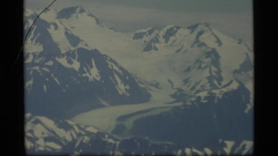 1977 Alaska 8mm aerial view of snow-covered peaks, dark ridges, and a glacier under a pale sky.