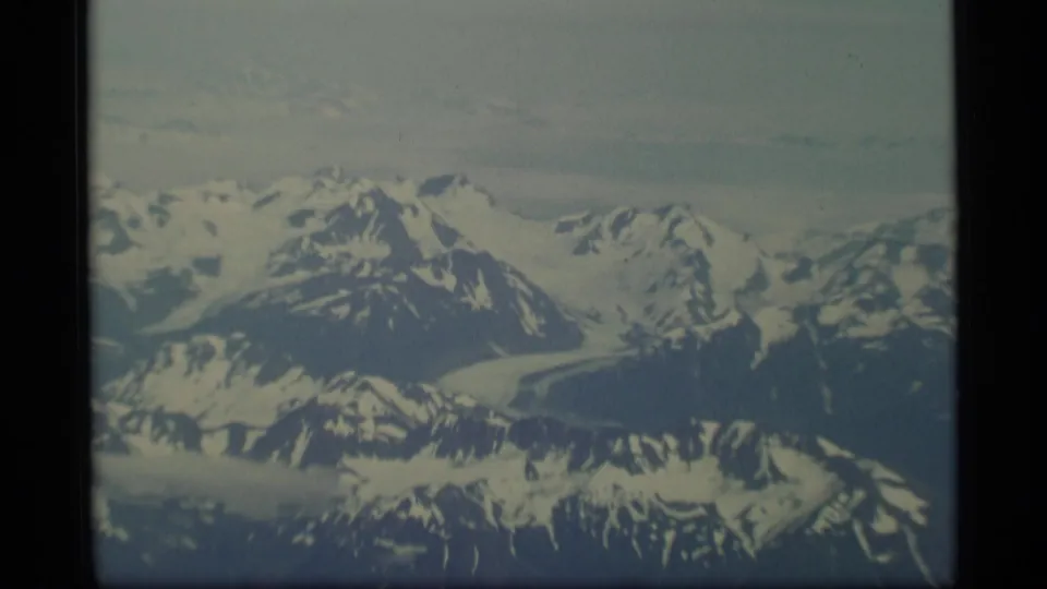 1977 Alaska aerial frame showing distant snowfields, glacier channels, and rugged mountain ridges.
