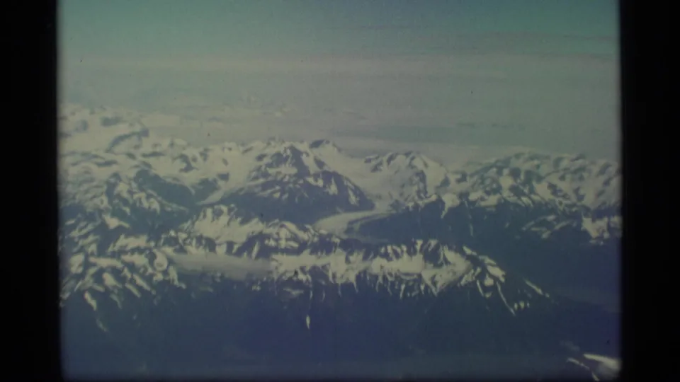 1977 Alaska 8mm aerial view across a wide snowy mountain range with a low cloud band near the horizon.