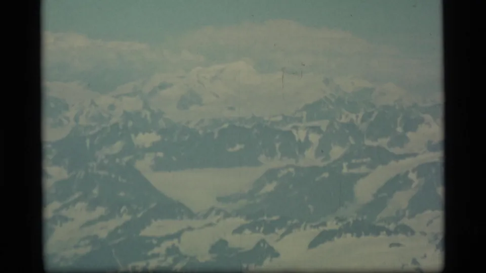 Faded 1977 Alaska aerial frame with snowy peaks, glacier basins, and soft blue-green 8mm color.